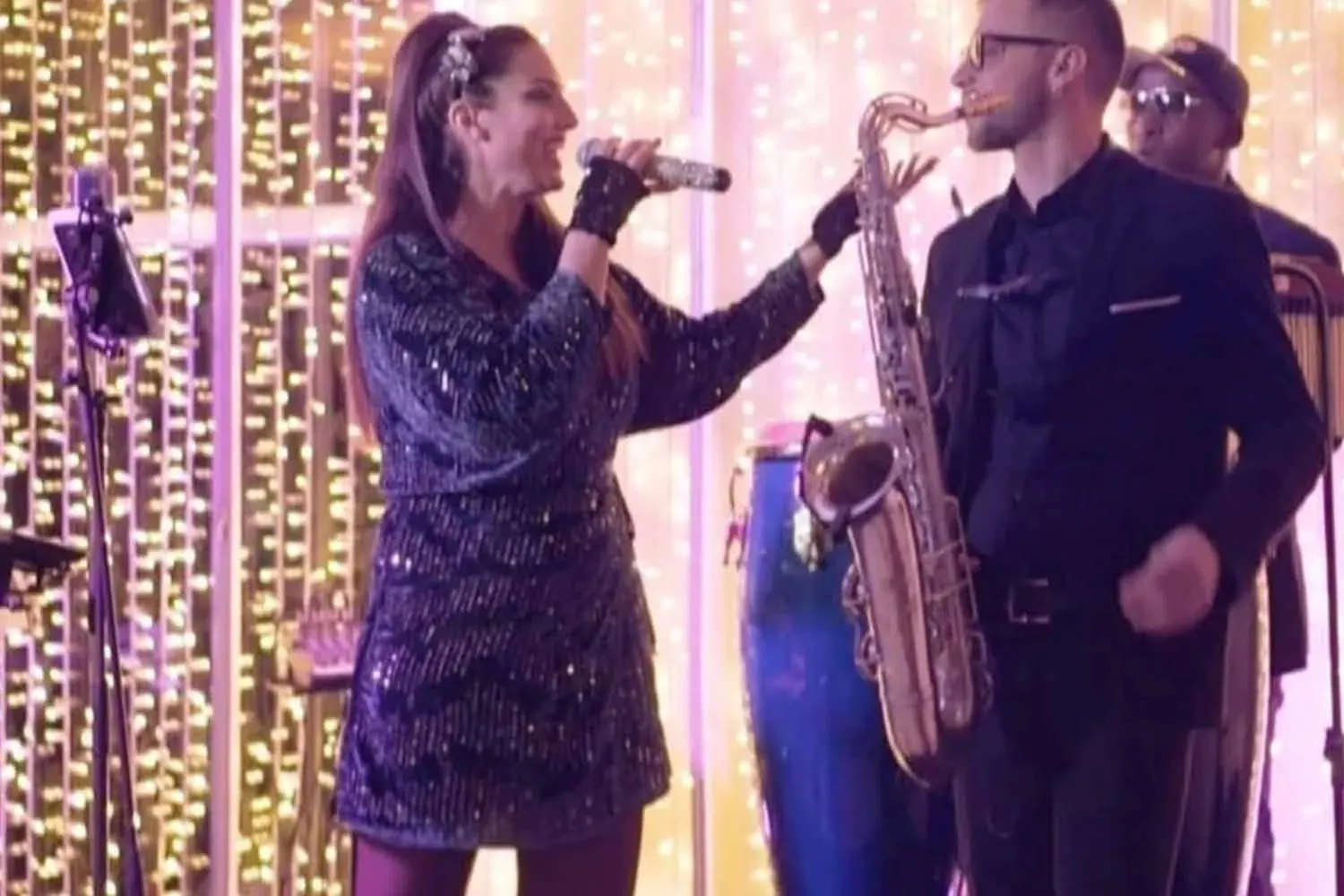 Chantal canta con il microfono in mano accanto al sassofonista, davanti a una tendina illuminata di luce calda.