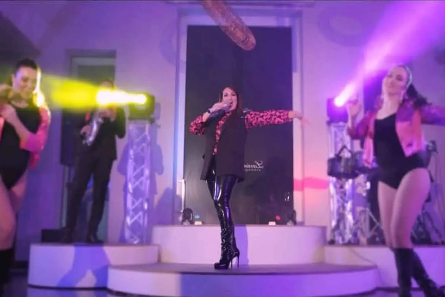 Chantal sul palco durante un dinner show: le ballerine ai lati, il sassofonista in primo piano e il fondale illuminato di viola.