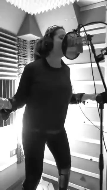 Chantal canta in cabina di registrazione, con le cuffie e il microfono da studio davanti a lei.