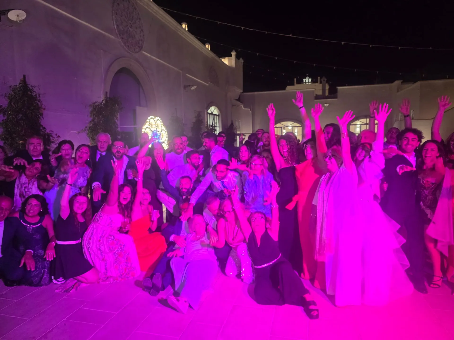 Una folla di invitati balla di sera nella corte di una masseria, sotto luci rosa.